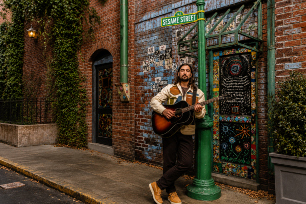 Noah Kahan holds an acoustic guitar while leaning against a green lamppost beneath a "Sesame Street" sign, in front of a mosaic-decorated brick wall, promoting his 2027 stadium tour.