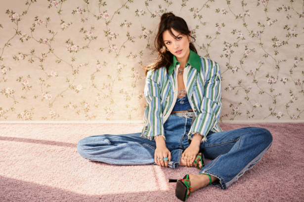 Olivia Rodrigo sits on pink carpet against floral wallpaper in a striped jacket and jeans, in press art around the release of her song 'Serena Joy.'