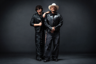 Pepe Walker and Gabito Ballesteros pose in coordinated black leather looks against a dark backdrop, one in sunglasses and the other in a white cowboy hat, promoting their single "A La Luz De La Luna."