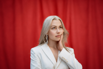 Phoebe Bridgers, whose album 'Lost Weekend' arrives Aug. 14, poses in a white blazer against a red curtain in a press portrait.
