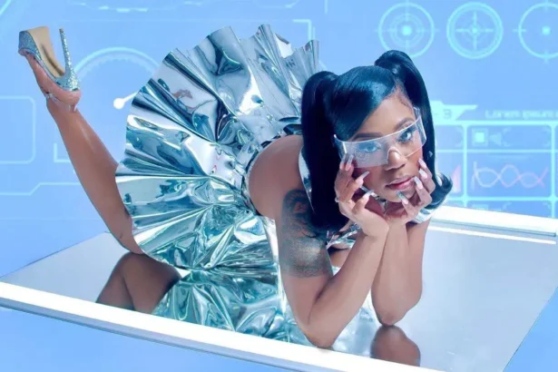 Rakiyah reclines on a mirrored surface in a metallic silver dress and futuristic visor, set against a blue sci-fi interface background, for her album 'My Collection of Boy Tears.'