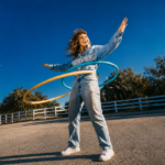 Remi Wolf laughs mid-motion in an all-denim outfit, playing with hula hoops outdoors against a bright blue sky, promoting her new album 'Mud.'