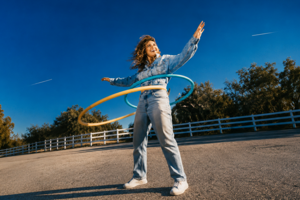 Remi Wolf laughs mid-motion in an all-denim outfit, playing with hula hoops outdoors against a bright blue sky, promoting her new album 'Mud.'