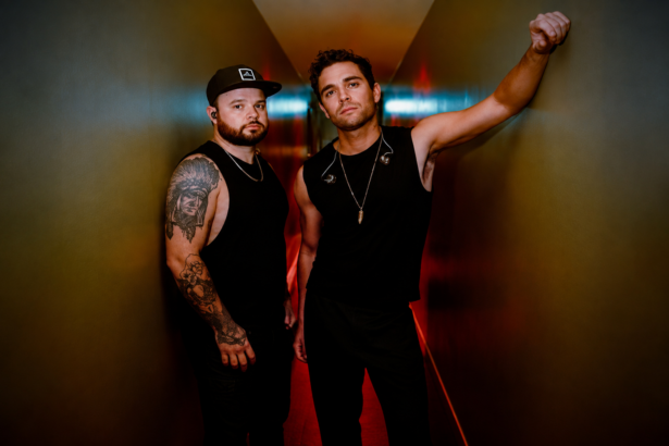 The two members of Royal Blood pose in black outfits in a moody, warmly lit corridor, in press art for their fifth album 'Dead Company' and single 'Ten Over Ten.'