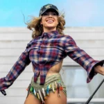 Slayyyter performs live in a plaid flannel shirt, embellished cap, and fishing-lure-adorned shorts, arms outstretched and smiling, during her 2026 tour.
