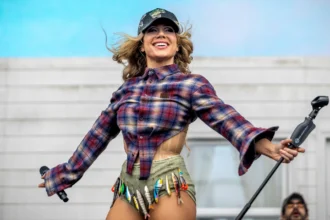Slayyyter performs live in a plaid flannel shirt, embellished cap, and fishing-lure-adorned shorts, arms outstretched and smiling, during her 2026 tour.