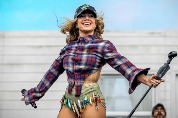 Slayyyter performs live in a plaid flannel shirt, embellished cap, and fishing-lure-adorned shorts, arms outstretched and smiling, during her 2026 tour.