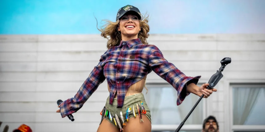 Slayyyter performs live in a plaid flannel shirt, embellished cap, and fishing-lure-adorned shorts, arms outstretched and smiling, during her 2026 tour.
