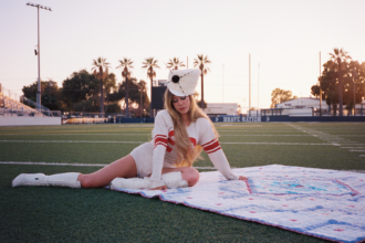 Slayyyter poses on a quilt on an empty football field at dusk in a vintage jersey and boots, from the "brand new chanel$" video.