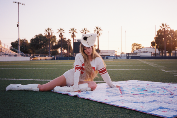 Slayyyter poses on a quilt on an empty football field at dusk in a vintage jersey and boots, from the "brand new chanel$" video.