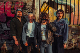 The four members of Bristol band Spin Class pose in denim and leather against a graffiti-covered wall at golden hour, in press art for their single 'Rip 'N' Dip.'