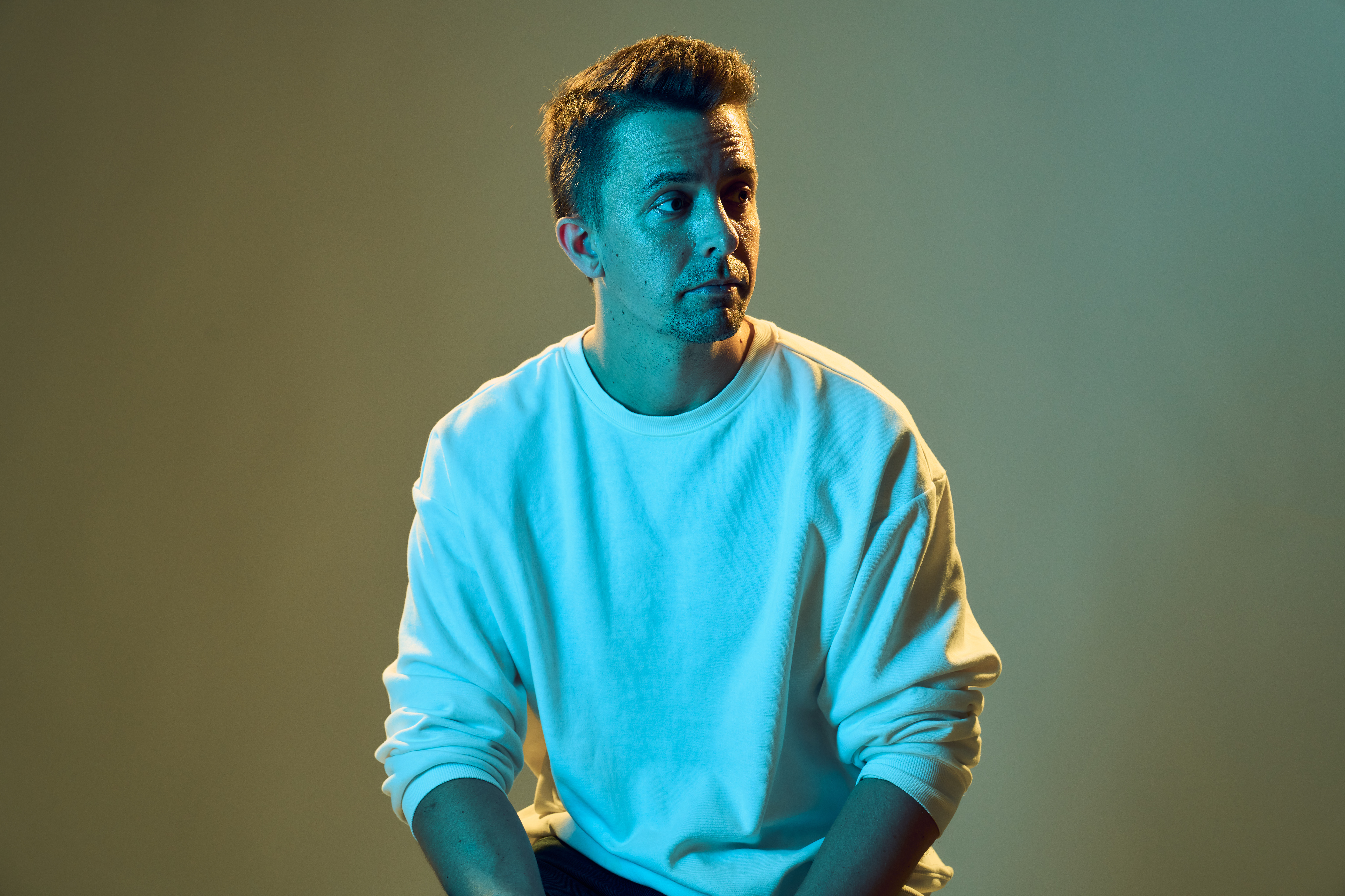 Steve Stinson posing in a white sweatshirt under cinematic blue and warm lighting against a muted green background.
