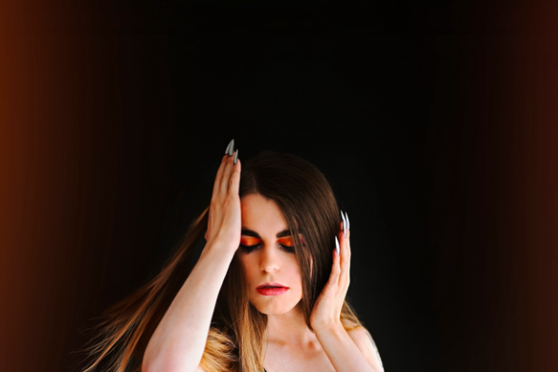Tom Ymani posing in a black glossy dress against a dark background, with long brown hair, dramatic eye makeup, and her hands raised near her face.