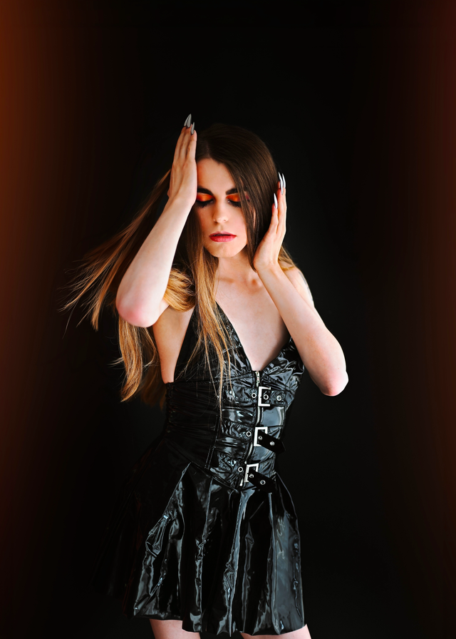 Tom Ymani posing in a black glossy dress against a dark background, with long brown hair, dramatic eye makeup, and her hands raised near her face.