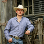 Country singer Tristan Roberson leans against a weathered wooden fence in a white cowboy hat, blue checked shirt, jeans, and a longhorn belt buckle, promoting his album 'Down For The Ride.'