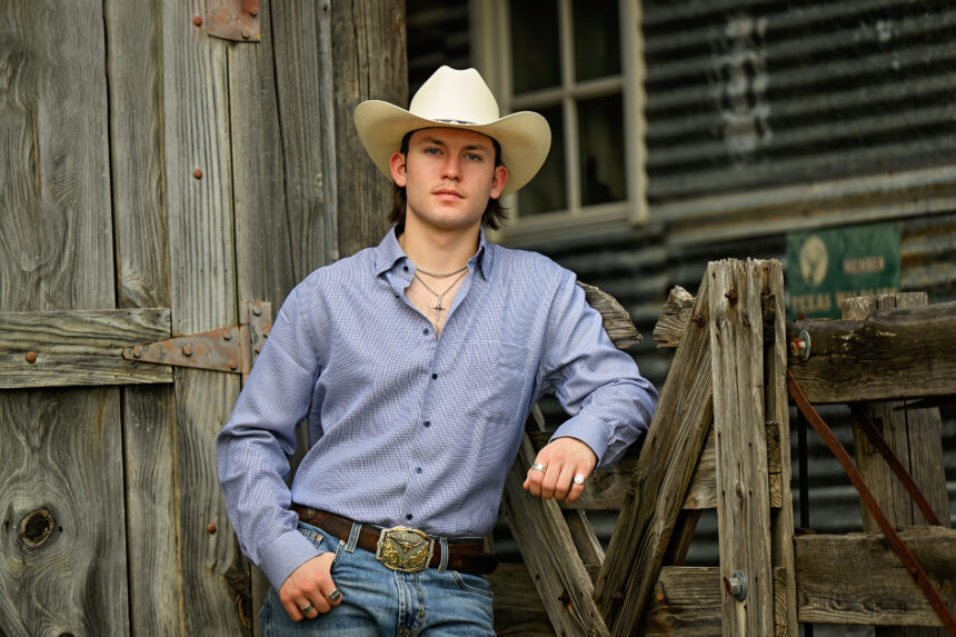 Country singer Tristan Roberson leans against a weathered wooden fence in a white cowboy hat, blue checked shirt, jeans, and a longhorn belt buckle, promoting his album 'Down For The Ride.'