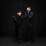 Twenty One Pilots' Tyler Joseph and Josh Dun pose against a black backdrop in dark clothing, one with red-streaked hair and red eye makeup, the other pressing black-painted hands to his face.
