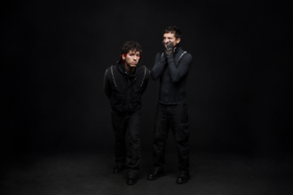 Twenty One Pilots' Tyler Joseph and Josh Dun pose against a black backdrop in dark clothing, one with red-streaked hair and red eye makeup, the other pressing black-painted hands to his face.