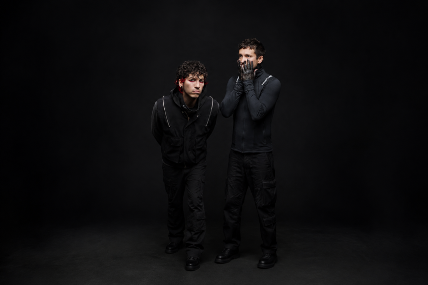 Twenty One Pilots' Tyler Joseph and Josh Dun pose against a black backdrop in dark clothing, one with red-streaked hair and red eye makeup, the other pressing black-painted hands to his face.