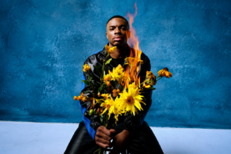 Vince Staples sits against a blue backdrop holding a bouquet of yellow flowers that is on fire, in press art tied to his 'Cry Baby' album and 2027 Australian tour.