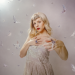 AURORA poses in a white lace top with red lipstick against a soft backdrop scattered with butterflies, in press art tied to 'All My Demons Greeting Me as a Friend (The Moth Tapes).'