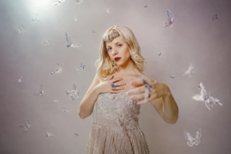 AURORA poses in a white lace top with red lipstick against a soft backdrop scattered with butterflies, in press art tied to 'All My Demons Greeting Me as a Friend (The Moth Tapes).'
