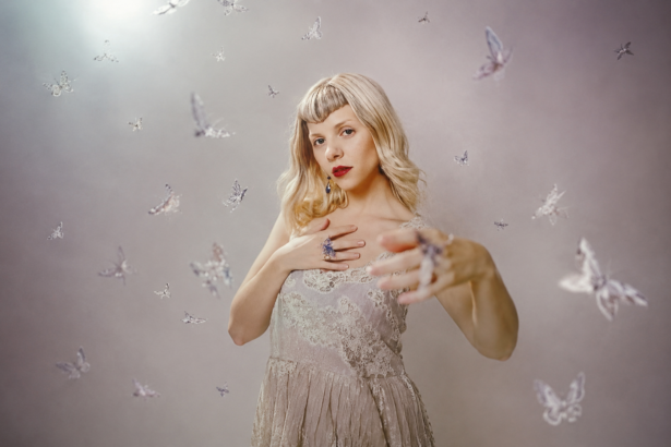 AURORA poses in a white lace top with red lipstick against a soft backdrop scattered with butterflies, in press art tied to 'All My Demons Greeting Me as a Friend (The Moth Tapes).'