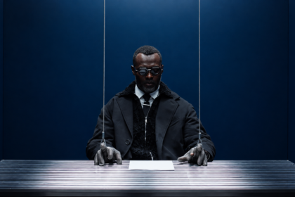 Actress at a metal desk in a blue room, his chainmail-gloved hands attached to wires like a marionette