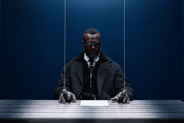 Actress at a metal desk in a blue room, his chainmail-gloved hands attached to wires like a marionette
