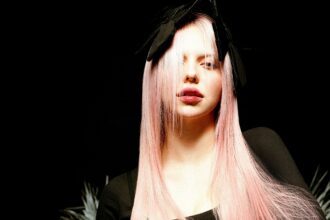 Adéla poses against a black background with long straight pink hair falling over one eye, wearing a black bow headpiece and dark top, her expression cool and direct in dramatic lighting.