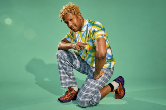 Mykki Blanco kneels against a mint green background with blond dreadlocks and face tattoos, wearing a colorful tie-dye polo shirt, blue plaid trousers, and orange-and-purple sneakers.