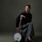 Rhiannon Giddens sits against a dark green backdrop holding a banjo, wearing a black sweater and checkered skirt with red flowers in her hair.