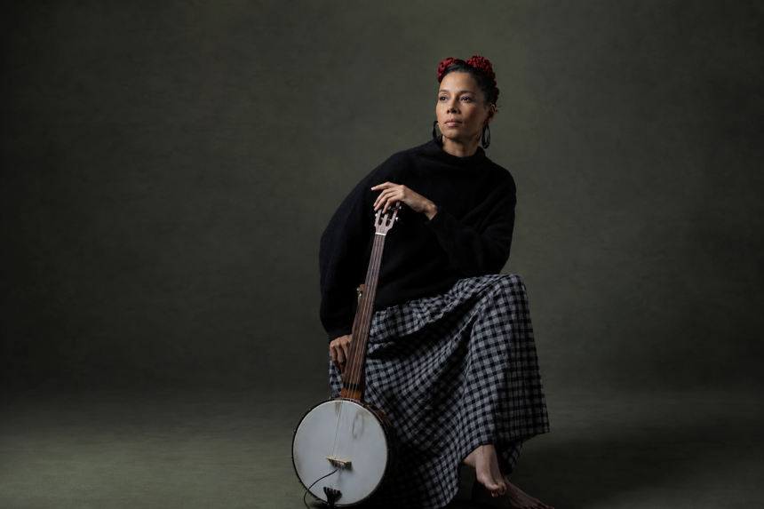 Rhiannon Giddens sits against a dark green backdrop holding a banjo, wearing a black sweater and checkered skirt with red flowers in her hair.