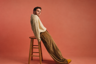 Alex Warren leans back against a wooden stool in a cream textured sweater, tan corduroy trousers, and yellow shoes, set against a warm terracotta background.