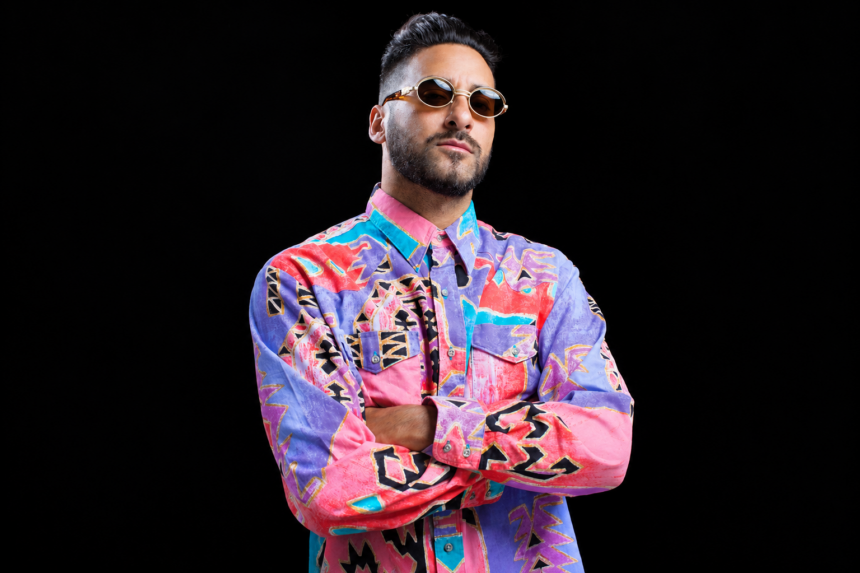 Armand Van Helden, who headlines New Year's Eve at Sydney's The Works, poses with arms crossed against a black background, wearing round tinted sunglasses and a vibrant pink, purple and teal patterned shirt.