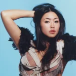 Beabadoobee posing with one hand in her hair, wearing a satin top with black feather shoulders against a pale blue backdrop