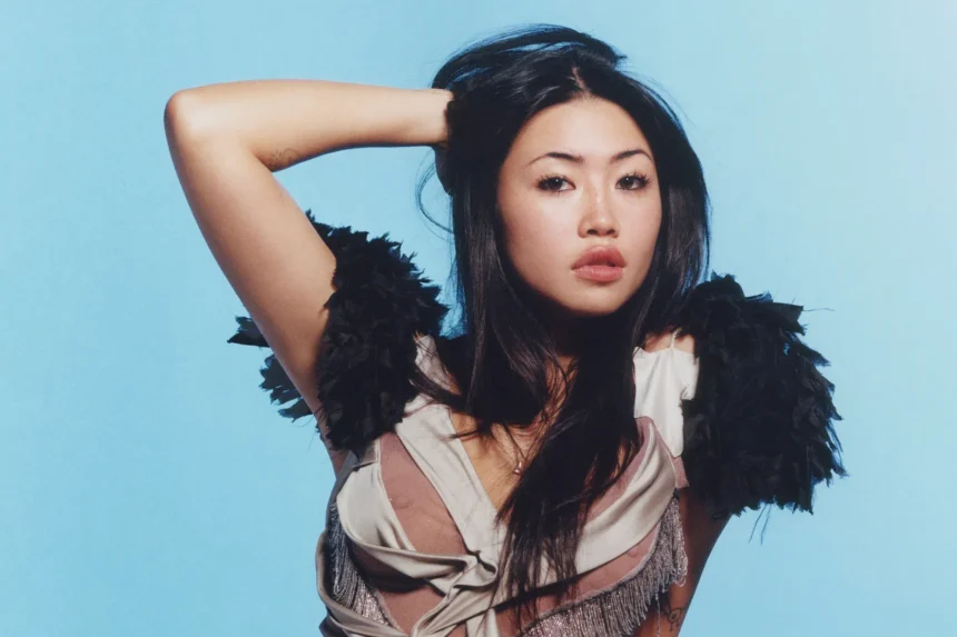 Beabadoobee posing with one hand in her hair, wearing a satin top with black feather shoulders against a pale blue backdrop