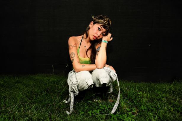 Beabadoobee crouches on grass against a dark wall in a green top and light jeans with visible tattoos, in press art tied to her 2027 Australian arena tour and album 'Pylon.'