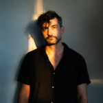 Bonobo (Simon Green) stands against a teal-lit wall in a dark short-sleeved shirt, his face half-lit by warm golden light in a moody portrait.