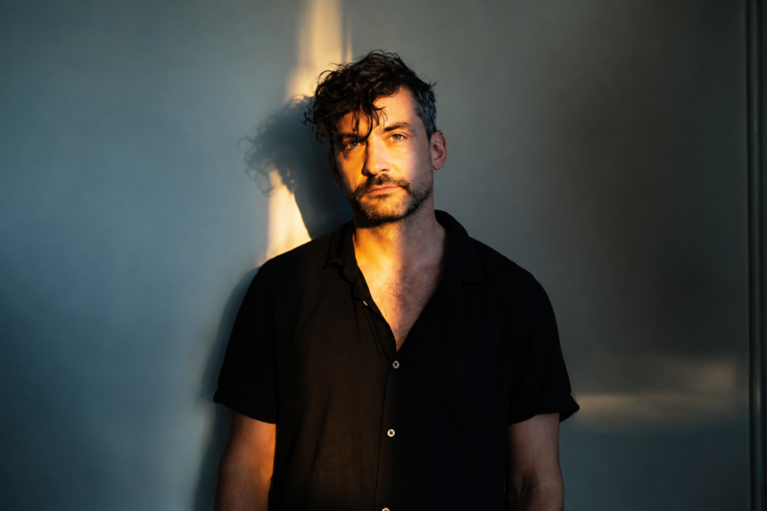 Bonobo (Simon Green) stands against a teal-lit wall in a dark short-sleeved shirt, his face half-lit by warm golden light in a moody portrait.