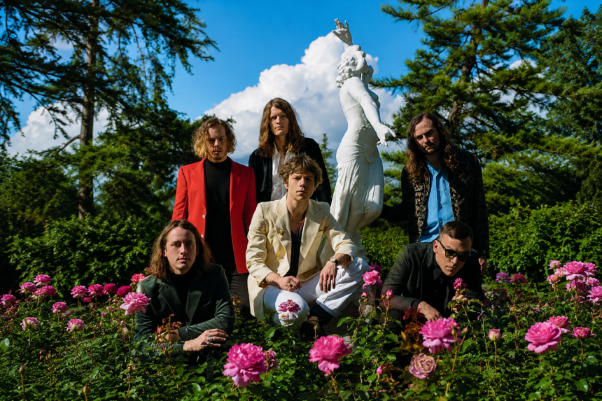 Cage The Elephant, who announced their 2027 'Dying In Reverse' tour, pose among pink roses in front of a white classical statue and blue sky, frontman Matt Shultz crouching center in a cream suit surrounded by the band in tailored jackets.