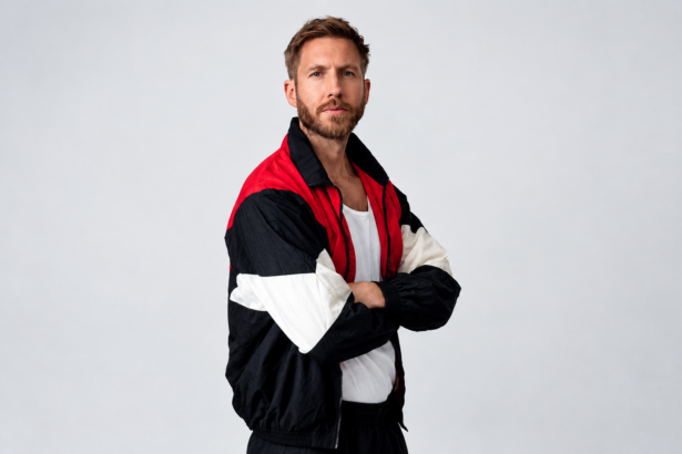 Calvin Harris stands with arms crossed against a plain gray backdrop, wearing a red, black, and white color-block track jacket over a white top.