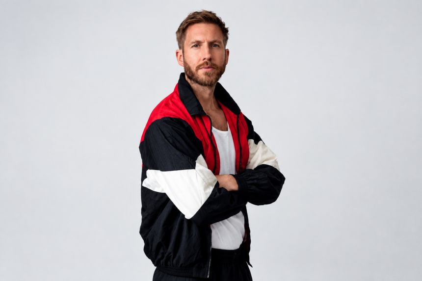 Calvin Harris stands with arms crossed against a plain gray backdrop, wearing a red, black, and white color-block track jacket over a white top.