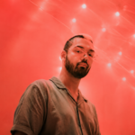 DJRUM stands in glasses and a collared shirt against a red-lit backdrop scattered with string lights, in press art tied to his 'DJ-Kicks' mix.