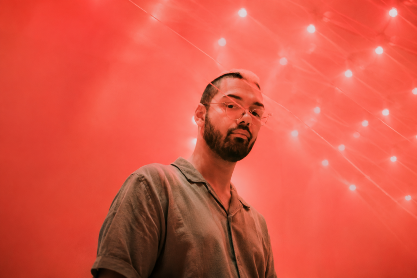 DJRUM stands in glasses and a collared shirt against a red-lit backdrop scattered with string lights, in press art tied to his 'DJ-Kicks' mix.
