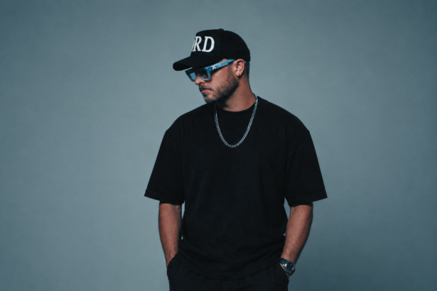 A house producer poses against a gray backdrop in an all-black outfit with a black cap, blue-tinted sunglasses, and a silver chain, looking to the side with hands in pockets.