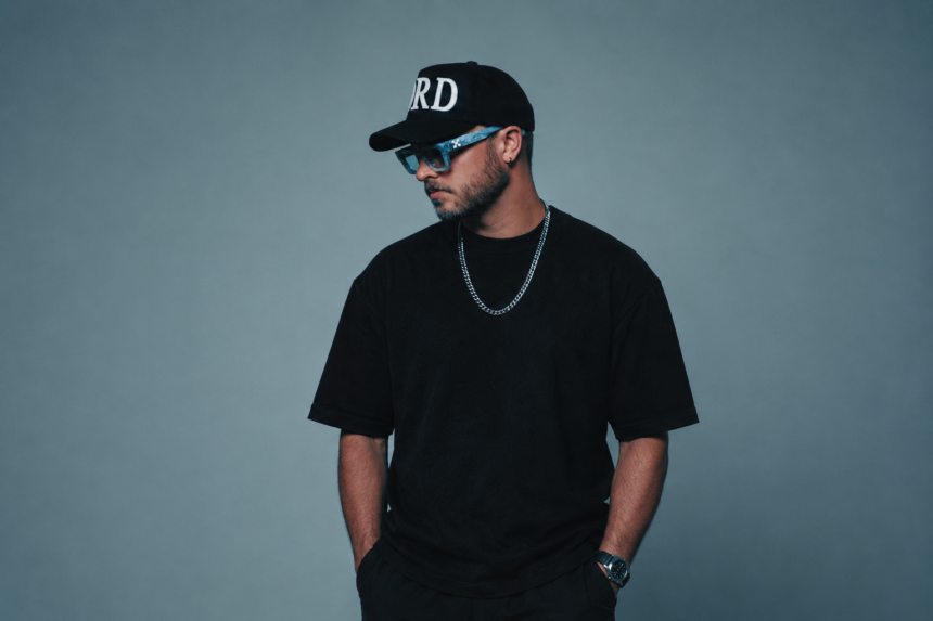 A house producer poses against a gray backdrop in an all-black outfit with a black cap, blue-tinted sunglasses, and a silver chain, looking to the side with hands in pockets.