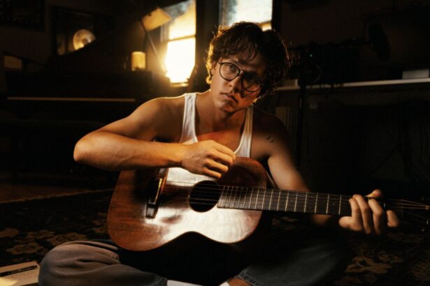David Kushner sits cross-legged on a patterned rug playing an acoustic guitar in warm, dim light, wearing a white tank top and round glasses with a notebook beside him.