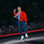 Harry Styles leaps mid-air while singing into a microphone on stage, wearing a red satin jacket, blue trousers, and white socks, with a packed crowd holding up phones behind him.