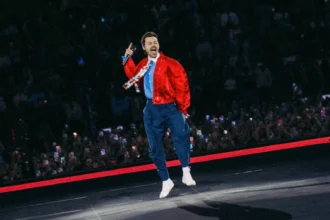 Harry Styles leaps mid-air while singing into a microphone on stage, wearing a red satin jacket, blue trousers, and white socks, with a packed crowd holding up phones behind him.